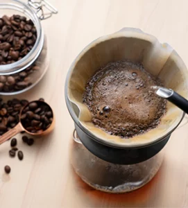 An overhead shot of water being poured from a gooseneck kettle into a filter filled with coffee grounds, showing the "bloom" effect, with a jar of whole beans and a copper scoop on a wooden surface.