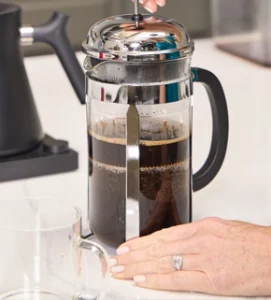 A close-up shot of a person’s hand pressing down the plunger of a stainless steel and glass French press filled with dark, rich coffee, set on a white marble countertop.