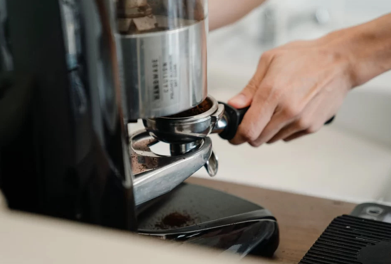 A close-up action shot of a professional coffee grinder dispensing freshly ground coffee into a portafilter held by a person’s hand.