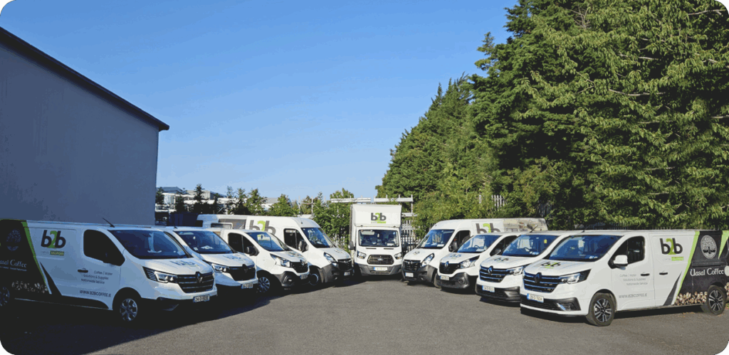 A wide shot of ten white service vans and a box truck with "B2B Coffee" and "Uasal Coffee" branding parked in a semi-circle in a paved lot.