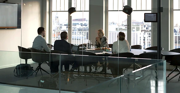 A wide shot of a glass-walled modern boardroom where four professionals are gathered around a dark oval table for a meeting, with large windows overlooking a city skyline.